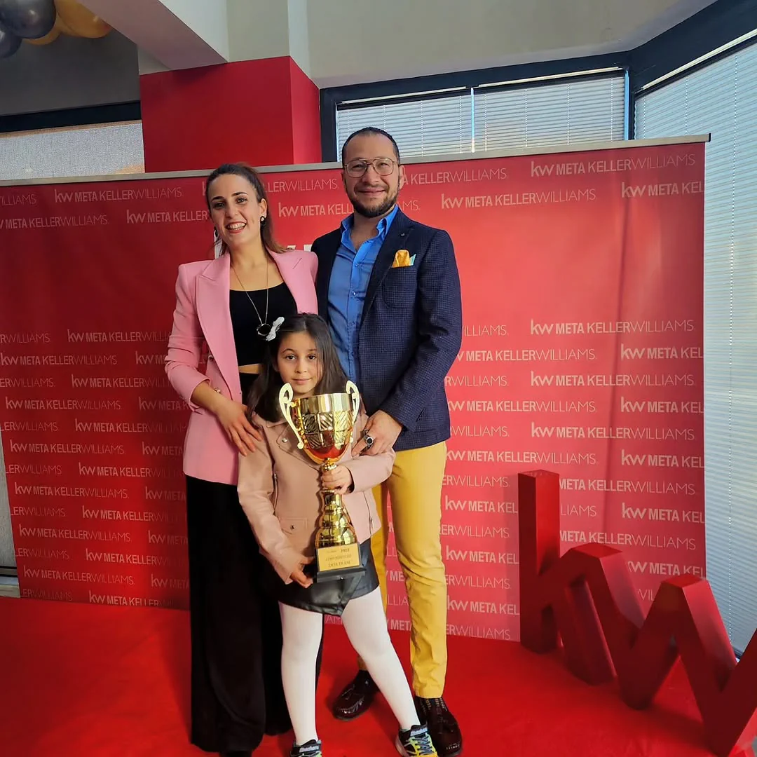 Pelin ve Serkan Alkan, kupayı tutan küçük bir kızla KW Meta panosunun önünde